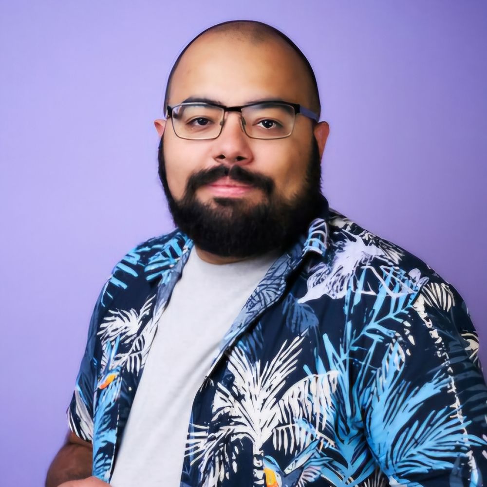 Bearded man with glasses wearing a colourful dark blue shirt with lead patterns in light blue and white across it, white t-shirt underneath this. Background of the image is purple